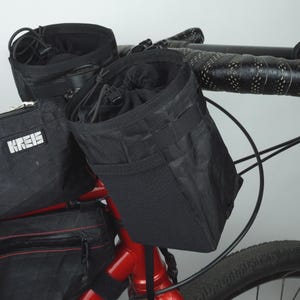 Puede incluir: Bolsas de manillar y bolsa de cuadro negras fijadas a un cuadro de bicicleta rojo. Las bolsas tienen un cierre de cordón y la bolsa de cuadro tiene una cremallera. El manillar tiene cinta negra.