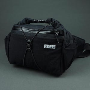 May include: Black fanny pack with the word "KREIS" in white block letters. The bag has a textured front panel, a zippered pocket, and a black cord detail. It features a top handle and a black strap.
