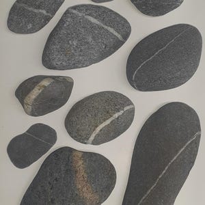 May include: A collection of ten smooth, gray and white stones with white lines running through them. The stones are arranged in a heart shape on a white background.