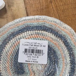 Puede incluir: Un rollo de tela con una mezcla de colores, incluyendo azul, melocotón y crema. La etiqueta dice "I Love Fall Most Of All" y "Jelly Roll 2.5" Strips". La tela está hecha en Corea.