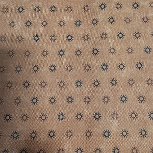 May include: Close-up of a tan fabric featuring a repeating pattern of small, stylised sunburst designs in dark blue and brown. The fabric has a textured appearance, suggesting a woven material.