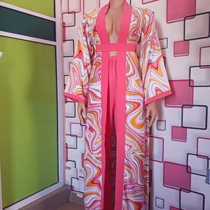 Puede incluir: Una blusa de manga larga rosa y blanca con un escote en V profundo y un pantalón a juego. La blusa tiene un patrón de remolino y un ribete rosa. El pantalón tiene una pierna recta.