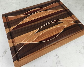 May include: Rectangular wooden cutting board with a juice groove. The board features a decorative design of curved lines in light and dark wood tones, including walnut, cherry, and maple. The board is approximately 18 inches long and 12 inches wide.