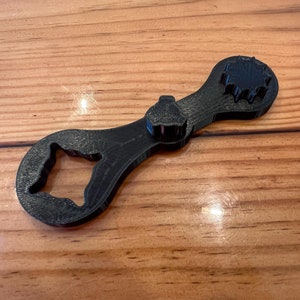 May include: A black plastic wrench with a unique design, featuring a star-shaped head and a curved handle with a cut-out.