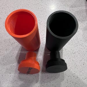 May include: Two cylindrical containers with lids. One container is orange and the other is black. Both containers have a screw-on lid.