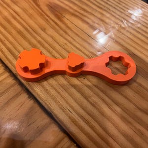 May include: An orange multi-tool with a gear-shaped end, a triangle-shaped end, and a star-shaped end. The tool is made of plastic and is lying on a wooden surface.