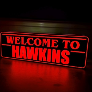 Puede incluir: Un letrero rectangular negro con las palabras "WELCOME TO HAWKINS" en letras rojas brillantes e iluminadas. El letrero es probablemente un artículo decorativo, posiblemente una luz nocturna o una decoración de pared.
