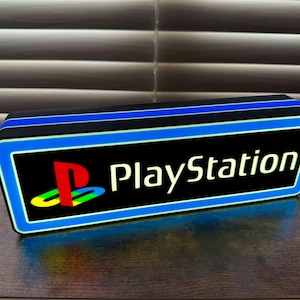 May include: A black and blue rectangular sign with a white border and a blue glow. The sign has the PlayStation logo and the word "PlayStation" in white letters.