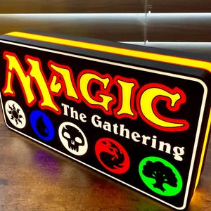 May include: A rectangular sign with the words "MAGIC The Gathering" in yellow and red lettering. The sign has a black background with various colored symbols and a yellow border.