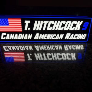 May include: A blue and white sign with the text "T. Hitchcock 9 Canadian American Racing" and an American flag.