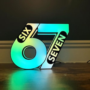 May include: Illuminated sign displaying the numbers "67" with the words "SIX" and "SEVEN" on the sides. The numbers are outlined in black and filled with a gradient of colors, including green, blue, and yellow. The sign is on a wooden surface.
