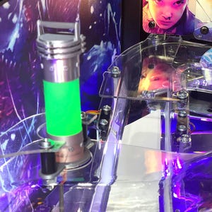 May include: A green glowing tube with a silver top and base, mounted on a pinball machine. The tube is surrounded by a clear plastic structure with screws and a purple light shining from below.