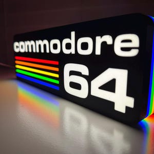 May include: Black rectangular sign with the words "commodore 64" in white. The sign has a rainbow stripe design and a rainbow-colored edge. The number "64" is in white.