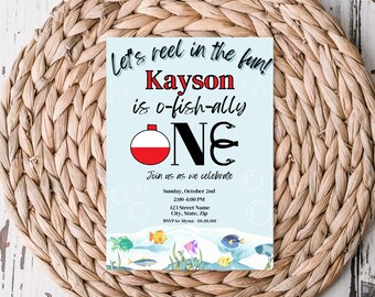 Peut inclure: Une invitation d'anniversaire bleue et blanche sur le thème de la pêche. L'invitation indique "Let's reel in the fun! Kayson is o-fish-ally ONE." Elle comprend un flotteur de pêche rouge et blanc et un hameçon. L'invitation indique également la date, l'heure et le lieu de la fête.