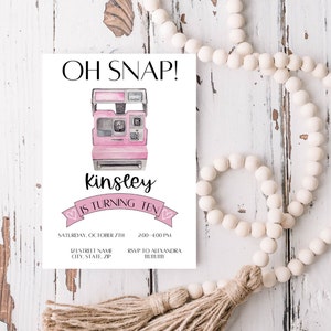 以下が含まれることがあります： ピンクと白の誕生日招待状で、ピンクのポラロイドカメラのイラストが描かれています。テキストには「Oh Snap! Kinsley is turning ten. Saturday, October 7th, 2:00-4:00 PM, 123 Street Name, City, State, Zip, RSVP to Alexandra, 11111111.」と書いてあります。