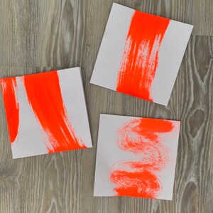 Può includere: Tre carte quadrate bianche con pennellate di colore arancione brillante. Le pennellate sono diverse su ogni carta. Una carta ha una singola pennellata verticale, un'altra ha una pennellata orizzontale e l'altra ha una pennellata ondulata.