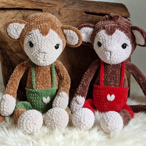 May include: Two handmade crochet monkey plush toys. One is brown with green overalls, the other is brown with red overalls. Both have white faces, ears, and feet. They are sitting on a white, fluffy surface.