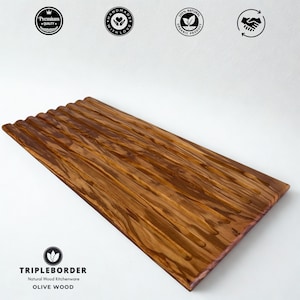 May include: A rectangular olive wood chopping board with a ridged surface. The board has a natural wood grain pattern and is branded with the text "TRIPLEBORDER Natural Wood Kitchenware OLIVE WOOD". The board is a premium quality, handmade, and organic product.