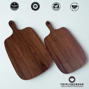 Puede incluir: Dos tablas de cortar de madera con asas. Las tablas son de color marrón oscuro con vetas de madera visibles. Las tablas están etiquetadas como "Premium Quality", "Handmade with Love" y "100% Natural Organic Product". También es visible la marca "TRIPLEBORDER Natural Wood Kitchenware".