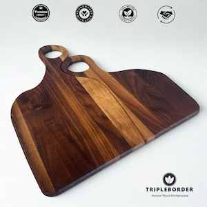 Puede incluir: Dos tablas de cortar de madera con acabado marrón oscuro. Cada tabla tiene un asa con una abertura circular. Las tablas están hechas de madera natural y presentan el texto "TRIPLEBORDER Natural Wood Kitchenware".
