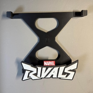 May include: Black wall-mounted display stand for Marvel Rivals. The stand has a unique hourglass-shaped design with the Marvel Rivals logo in white and red. The stand is made of black material and is designed to hold a gaming device.