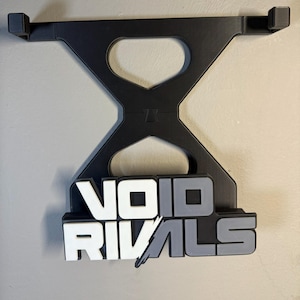 May include: Black wall-mounted gaming headset holder with the text "VOID RIVALS" in white, gray, and black. The holder has a unique geometric design with two arms to hold the headset.
