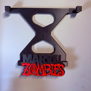 May include: Black wall-mounted shelf with the words "MARVEL ZOMBIES" in black and red. The shelf has a unique hourglass-like design with two rectangular supports at the top. The text is in a bold, stylized font.