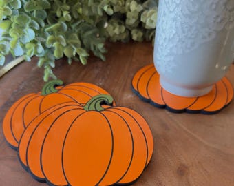May include: Orange pumpkin-shaped coasters with black outlines and green stems. A grey mug sits on one of the coasters. The coasters are on a wooden surface, with greenery in the background. The coasters are a fall-themed home decor item.