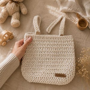 May include: A cream-colored, crocheted tote bag with two handles secured by wooden buttons. The bag has a rectangular shape and a small, brown leather tag. A wooden teddy bear, a wooden car, and a wooden scoop are also visible.