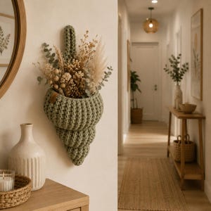 May include: A sage green, crochet wall planter shaped like a seashell, filled with dried flowers and foliage. The planter is mounted on a cream-colored wall, with a wooden table and mirror in the foreground.