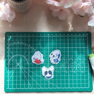 Könnte beinhalten: Drei gestanzte Vinyl-Aufkleber mit stilisierten Zeichentrickfiguren aus dem Spiel Hollow Knight, darunter eine weiße Figur mit rotem Umhang, eine blau-weiße Figur und ein kleines weißes Wesen, die alle auf einer grünen Schneidematte liegen.