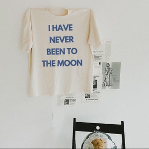 Puede incluir: Una camiseta beige con el texto "I have never been to the moon" impreso en azul.