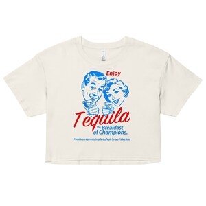 Può includere: Una maglietta bianca corta con un'illustrazione in stile vintage di un uomo e una donna che brindano con le bevande. Il testo "Enjoy Tequila The Breakfast of Champions." è stampato in rosso e blu.