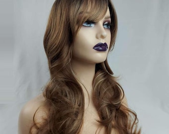 May include: A brown wig with long, wavy layers and a side-swept bang. The wig features dark roots that transition into lighter brown and blonde highlights. The wig is styled on a mannequin head with blue eyes and dark purple lipstick.
