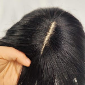 May include: Close-up of a black wig with a visible part. The hair is dark and sleek, with a natural-looking part that reveals the scalp. The wig appears to be made of synthetic fibers, and the overall style is simple and elegant.