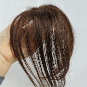 May include: A brown hairpiece with long, straight strands. The hairpiece is held up against a white background. The hair appears to be a medium brown colour with some lighter highlights. The hairpiece is likely a hair extension or wig.