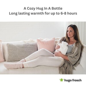 May include: A woman is sitting on a white couch holding a white hot water bottle with a gray and white cover. The hot water bottle is shaped like a bottle and has a long neck. The woman is wearing white pants and a gray sweater. The couch has a white throw pillow and a gray throw pillow. The text "A Cozy Hug In A Bottle" and "Long lasting warmth for up to 6-8 hours" is displayed above the image. The text "hugo frosch" is displayed below the image.