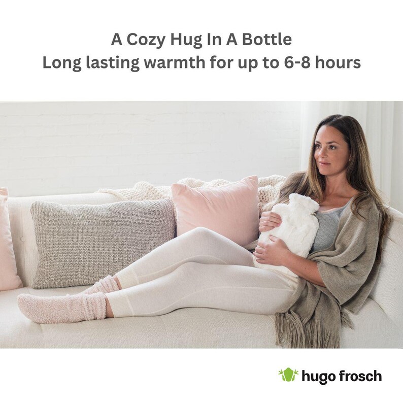 May include: A woman is sitting on a white couch holding a white hot water bottle with a gray and white cover. The hot water bottle is shaped like a bottle and has a long neck. The woman is wearing white pants and a gray sweater. The couch has a white throw pillow and a gray throw pillow. The text "A Cozy Hug In A Bottle" and "Long lasting warmth for up to 6-8 hours" is displayed above the image. The text "hugo frosch" is displayed below the image.
