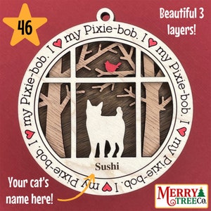 Puede incluir: Un adorno navideño de madera con la silueta de un gato en una escena de ventana. El adorno está en capas y tiene el texto "Me encanta mi Pixie-bob" repetido alrededor del borde. El nombre del gato "Sushi" está escrito debajo de la ventana.