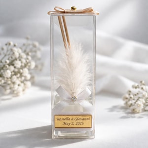 May include: A clear, rectangular favour box with a white feather, wooden sticks, and a white ribbon. The box has a gold label with the text "Rossella & Giovanni May 2, 2026". A small bow adorns the top.