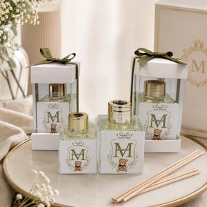 Op de afbeelding: Vier vierkante glazen geurverspreiderflesjes met gouden doppen, voorzien van een etiket met de letter M, een illustratie van een teddybeer en een bloemenkrans. Twee flesjes zitten in witte doosjes met olijfgroene lintstrikken en vier houten rietjes.