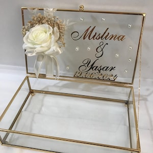 Może przedstawiać: Szklana skrzynka z pozłacanym wykończeniem i białą różą oraz suszonymi kwiatami na górze. Pokrywa ma wygrawerowane imiona "Mislina & Yasar" oraz datę "19.06.2024" złotymi literami.