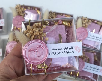 May include: Pink and gold gift boxes filled with a pink prayer rug, a string of pink beads, a small bouquet of dried flowers, and a pink wax seal with a footprint design. The boxes have a label with the Arabic text "اللهم انبتها نباتا حسناً وجعلها قرة عين لوالديها" which translates to "O Allah, make her grow up well and make her a comfort to her parents."