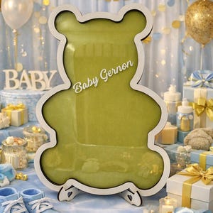 Puede incluir: Un marco de madera con forma de oso con contorno blanco e interior verde, ideal para un baby shower. El texto "Baby Gernon" se muestra en el marco. Zapatos de bebé azules y cajas de regalo en primer plano.