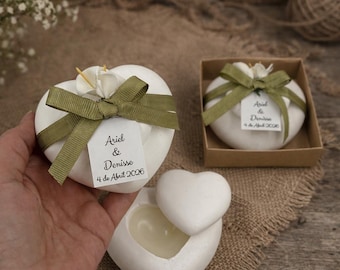 May include: White heart-shaped candle containers with lids, decorated with olive green ribbons and white tags printed with "Ariel & Denisse 4 de Abril 2026". One candle is open, one is closed with a ribbon, and one sits in a brown box.