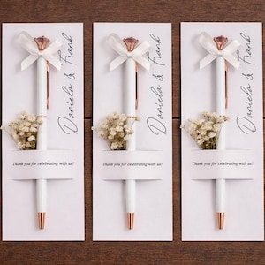 Puede incluir: Tres bolígrafos blancos con detalles en oro rosa, cada uno adornado con una parte superior en forma de diamante y un lazo blanco. Los bolígrafos se presentan en tarjetas blancas con los nombres "Daniela & Frank" y el mensaje "Thank you for celebrating with us!"