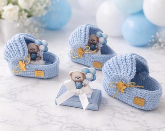 May include: Three light blue crocheted cradles and one rectangular light blue box. Each cradle features a small teddy bear holding blue and white balloons, a gold ribbon bow, and a gold rectangular tag. The box has a white ribbon and a teddy bear.