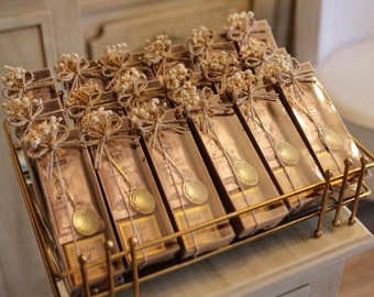 May include: A gold metal stand filled with small, clear boxes tied with twine and decorated with dried white flowers. Each box contains a small, gold-coloured spoon.
