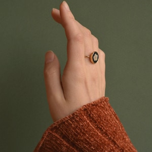 Puede incluir: Un anillo dorado con un centro ovalado negro con un diseño floral blanco. El anillo se lleva en una mano con una manga color óxido frente a un fondo verde. El diseño del anillo es simple y elegante.