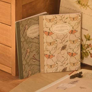 Puede incluir: Dos cuadernos de tapa blanda. El cuaderno de la izquierda tiene un fondo verde con pájaros marrones sobre ramas. El de la derecha tiene un fondo crema con mariposas naranjas y enredaderas verdes. Ambos tienen una etiqueta central que dice NOTEBOOK.